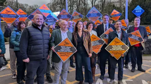 RYAN PRIEST Jane Dodds and 20 or so Lib Dem activists standing and smiling holding orange and brown placards saying "Welsh Liberal Democrats winning here" with English and Welsh language wording.