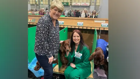 Laura Crombie Two women at Crufts. The one on the left has blonde hair and is wearing a floral shirt and jeans and is smiling at the camera. The second woman is sat down next to a dog in a booth, wearing a green suit.