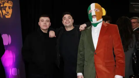 Getty Images Liam Óg Ó hAnnaidh, Naoise Ó Cairealláin and J.J. Ó Dochartaigh aka Mo Chara, Móglaà Bap and DJ Próvaà of Kneecap at the BAFTAS. Liam has short dark hair wearing black clothing. Naoise has dark wavy hair with his arms leant on the shoulders of Liam and JJ, he is wearing black clothing and rings. JJ is wearing a tri colour balaclava and tri colour suit. They are standing next to a BAFTAS sign.
