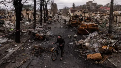 Getty Images A man pushes his bike through debris and destroyed Russian military vehicles on a street on April 06, 2022 in Bucha, Ukraine.