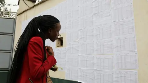 Reuters A woman wearing red with long braids looking for her name against an extensive list.