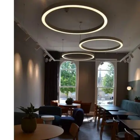 Brayford Creative Hub An empty room with high ceilings, chairs and tables. Two large windows are at the far end of the building and three large circular lights are hanging from the ceiling.