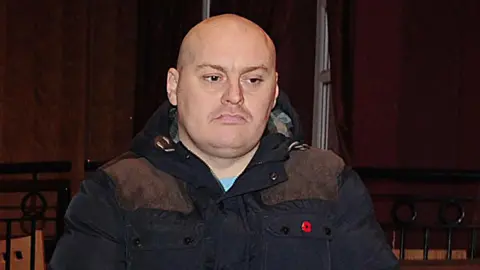 Ian Ogle is seen wearing a navy coat with a poppy pin.
He is bald, with a light stubble and frowning.
