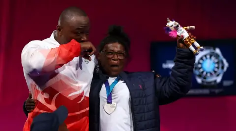 Person wearing glasses, a dark quilted jacket and a silver medal on a blue ribbon stands beside another person in a white and red sports jacket. The medal wearer raises a colourful mascot toy while standing on a sports event stage with a red backdrop.