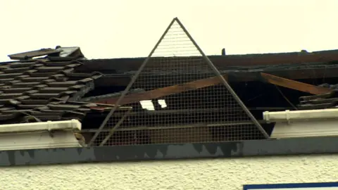 The roof of a building after a fire. Large parts of the roof is missing. The sky is grey and cloudy. 