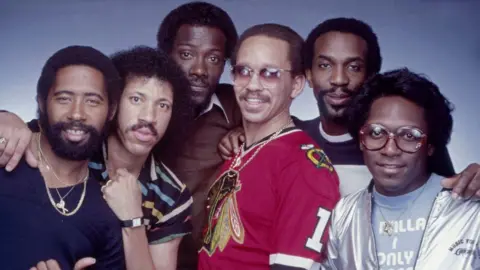 Getty Members of Commodores pose for a portrait in Los Angeles, California, in the 1980s. They are wearing a number of colourful outfits and all have their arms around each other.