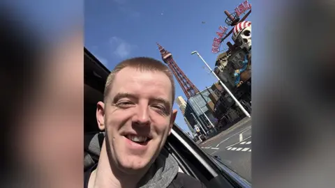 Dazza Darren Dowling (left) with short brown hair wearing a black t-shirt and grey hoodie with Blackpool Tower and Coral Island amusements in the background. He is smiling. 