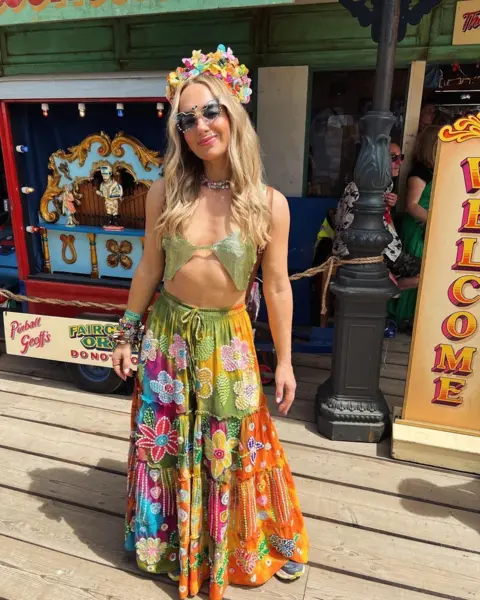 Sophie Ackers Sophie Ackers stands in front of a pier-style attraction at Glastonbury. She has long blonde hair and wears a flower crown and sunglasses. She wears a green bikini-style top and a floor-length multicoloured skirt.