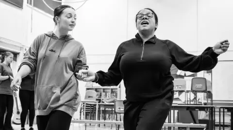 Cat Humphries Two female performers in rehearsal. One woman is looking to her dance partner on her right, and is wearing a hoddie. The other woman has her mouth open, singing, with her right arm out. There are two more women in the background. 