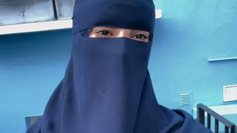 Salsabil in a blue niqab and headscarf, standing in front of a blue wall.