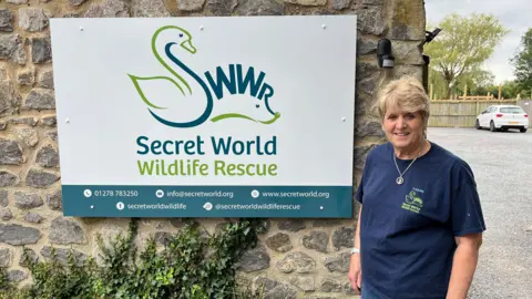 Pauline Kidner, wearing blue t-shirt, in front of Secret World Wildlife Rescue sign that is attached to brick wall.