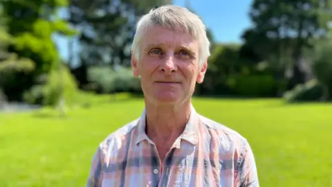 Jeff has short well kept silver/grey hair. He is wearing a salmon pink checked shirt.