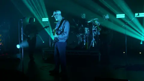 The Snuts perform live on stage. Jack the vocalist/guitarist is clearly visible as he stands in front wearing a light cap, a light jacket and jeans. The other band members are standing behind and aren't so clearly visible. The lights on stage are turquoise.