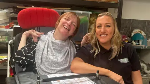 Vicky, in her electric wheelchair smiling at the camera with one hand around her sister Sarah, who is sat in a chair next to hair