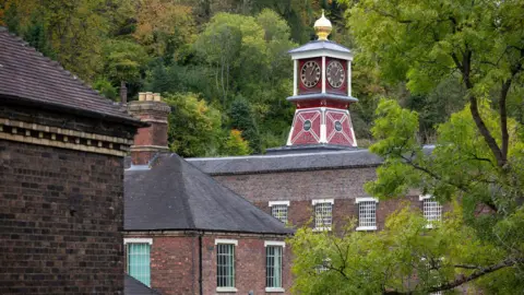 National Trust A brown brick building with black roof tiles and white windows with latticing. There is a red clock tower on top of the roof with a gold and black clock face. On top is a gold point.