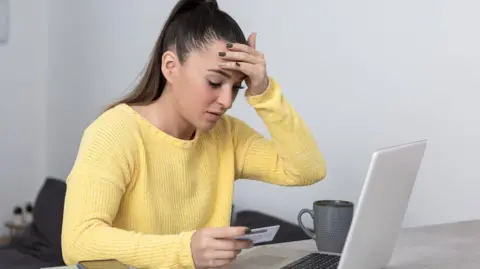 Stressed woman in yellow jumper trying to use bank card on her laptop