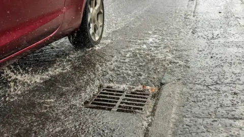 A red car travels past a flooded drain cover on a road.