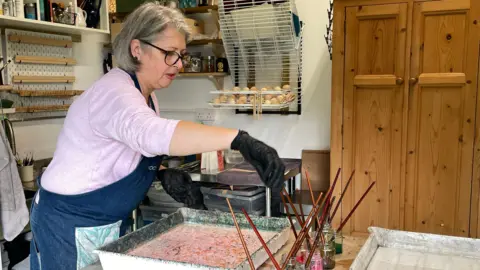 A woman wearing dark blue overalls in a art studio, over a water bathe with a number of jars holding paint brushes on the same table