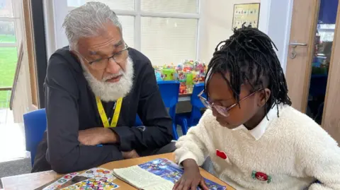 BBC An adult and a child sit at a classroom table with books and learning materials. The child is reading from an open book while another book lies nearby. Colourful educational posters and toys are visible in the background, and a door with a notice is on the right. A yellow volunteer lanyard and leaflets about reading support are on the table.