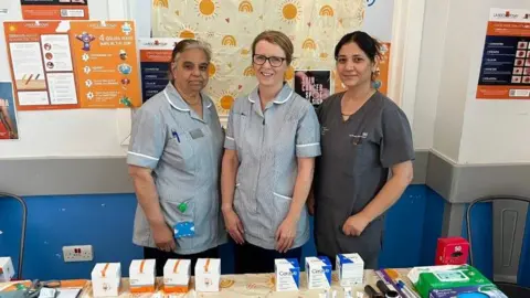Three women in medical scrubs smile at the camera while standing next to each other. In front of them is a table with sun cream and SPF products on it.