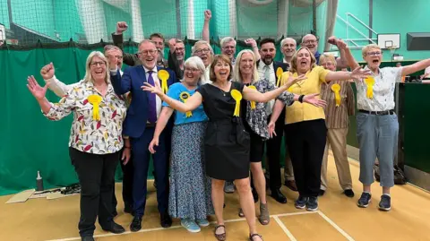 Liberal Democrat councillors cheering in a school gymnasium. Some of them have their arms outstretcehd in celebration. They are all wearing yellow rosettes on their cheer
