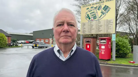 ELLEN KNIGHT/BBC Howard pictured looking into the camera with a serious expression. He's wearing a white collared shirt with thin blue stripes, and a V-neck navy blue woollen jumper over the top. He's stood outside, in front of a large yellow sign that reads 'Atcham Business Park' in dark green writing, with a map of the site laid out beneath the name. Below the sign are two red postboxes, and beyond it two large, green warehouses can be seen. There are trees and shrubbery planted on grass verges either side of Howard, and the tarmac ground is slick with recent rain. The sky is grey and overcast. 
