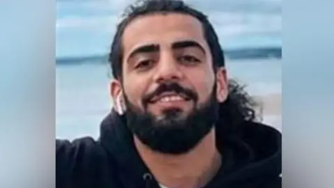 Zherko Rasul Osman has long black hair tied back, and a dark beard. He is wearing a black hoodie and has a earphone in his ear.