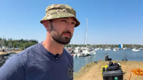 Chris is looking off to the right side of the image. He is wearing a hat which is covered in camo print. He is wearing a blue t-shirt. Behind him is Beaulieu river where the divers are in the water. There are boxes of diving equipment to the bottom right of the picture.