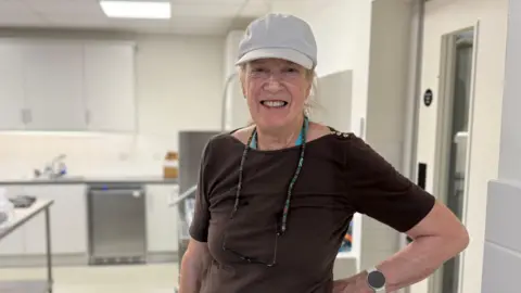 Jamie Niblock/BBC Dilys Hall wearing a cap and a brown top standing in a kitchen area. She is smiling broadly at the camera. 