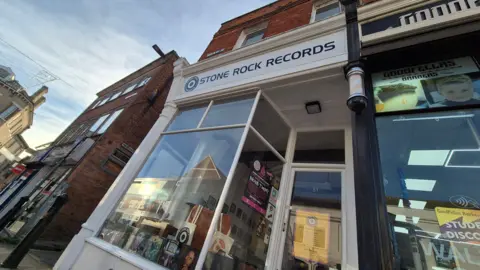 Jamie Niblock/BBC The outside of a record shop, with a white front, and posters and records in the window. Other shops are either side of it. 