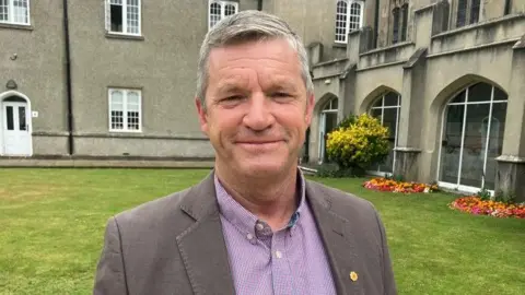 Brian Davies is smiling at the camera. He has short grey hair and is wearing a pink and blue chequered shirt and a light brown jacket.