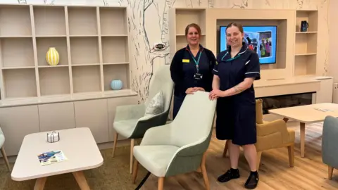 Severn Hospice Two women with brown hair are smiling and standing in a room, wearing navy skirts and t-shirts. There are two sets of tables and chairs and a television mounted onto the wall. On the left there are shelves built into the wall, with a tree painted behind them