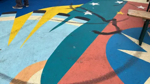 A colourful mural which is painted on the floor has dark black tyre marks over large sections of it.
