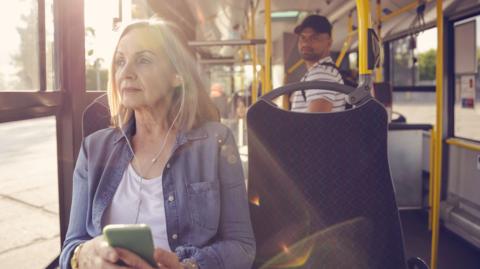 Elderly female is looking away while traveling in public transport on sunny day. She is wearing denim shirt.