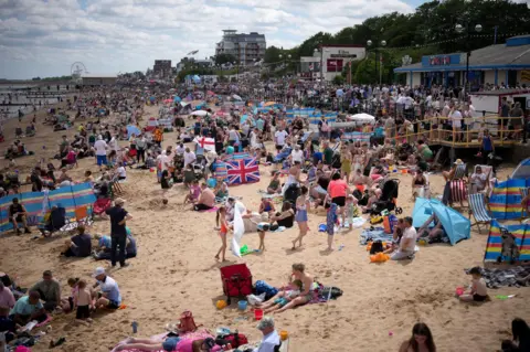 Getty Images A golden beach packed with people stretching far into the distance on a summer's day. Colourful wind-breakers and flags can be seen. To the left are seaside hut-type buildings and trees.