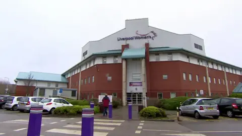Exterior shot of Liverpool Women's Hospital, the main building is brown/orange, the roof is white. Liverpool Women's is written on the front in purple. Purple bollards lead up to the entrance.