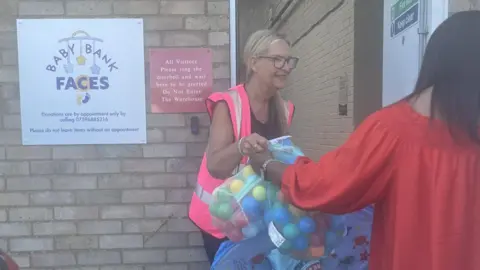 Bedford BabyBank A bag of balls, being given to a woman, in a pink high-vis jacket, by an open door in a brick building. You can see the back of a woman in a red dress. 