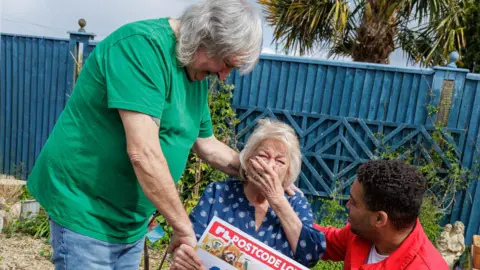 Postcode Lottery In a garden, a painted blue wooden fence behind. Lesley sits in the middle, overwhelmed holding a hand over her mouth and the other on a giant cheque. She wears a blue top. Philip stands next to her, in a green t-shirt and jeans. He comforts her with one hand on her shoulder. To the right, someone from the postcode lottery in a red shirt, also has his arm round her.