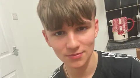 A selfie of a teenage boy who has a half-smile, with fair hair in a fringe cut straight across his forehead. He is wearing a black Nike top and appears to be in a kitchen, with a kettle, black tiles and white wall in the background.