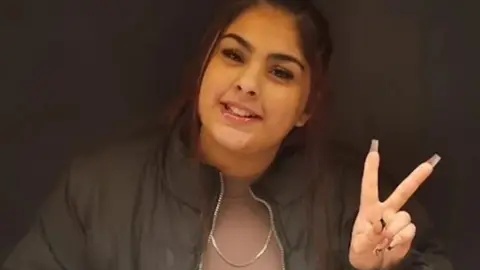 Aaliyah has dark hair in a ponytail, brown eyes and is smiling at the camera. She is wearing a dusty pink t-shirt with a black jacket over the top, she is holding up a peace sign with her fingers. 