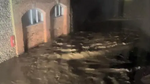 A river has burst its banks by Chard Junction in Somerset. Murky water is seen flowing down a road.