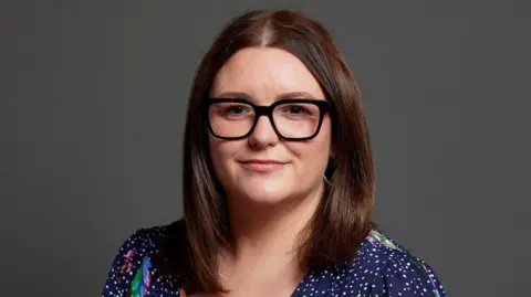 UK Parliament Leigh Ingham, a woman wearing a navy blue dress with white spots and a coloured pattern. She is pictured against a dark grey backdrop.