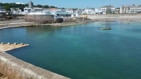 BBC Havre Des Pas bathing pool. The large tidal pool is full or turquoise water. There are steps leading up to an area used for seating and a cafe 