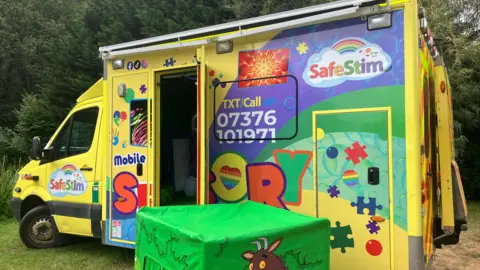 BBC A yellow ambulance which has been been transformed into a sensory van. On the side of the van there are colour stickers and symbols. The door of the van is open and there is a green box next to it. 