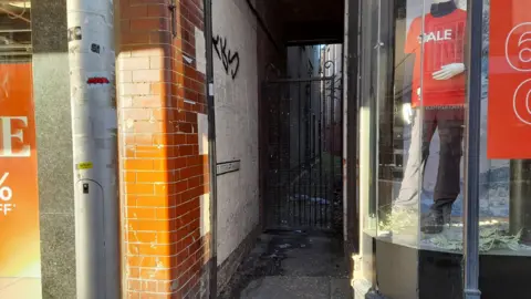 A black gated narrow passageway in-between two shops on a high street. The words 'St Peter's Passage' can be seen on the side of the wall.