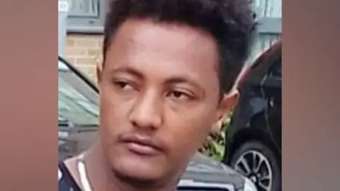 Adisu Chala is pictured outside a building in front of two vehicles. He is wearing a black and white top and necklace. 