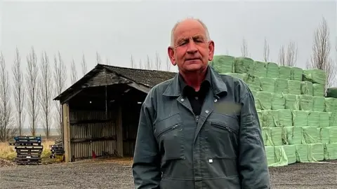 Cllr Julian Kirk wears dark blue overalls. He stands in front of a large barn and bales of animal forage wrapped in green plastic. 