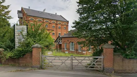 Google The outside of the building seen from the road. There is a wooden gate in the foreground and beyond it across a concrete area there are red brick buildings with windows up the side.