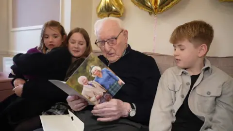Richard Knights/BBC Ron sitting on a sofa with three children. He is looking at a birthday car that has a picture of King Charles III and his wife on it.
