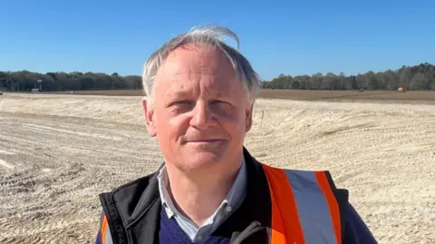 Richard Daniels/BBC A head and shoulders image of Andrew Blenkiron, who is wearing an orange high-vis over a black jacket, blue jumper and collared shirt. He is looking straight into the camera while standing on his farm.